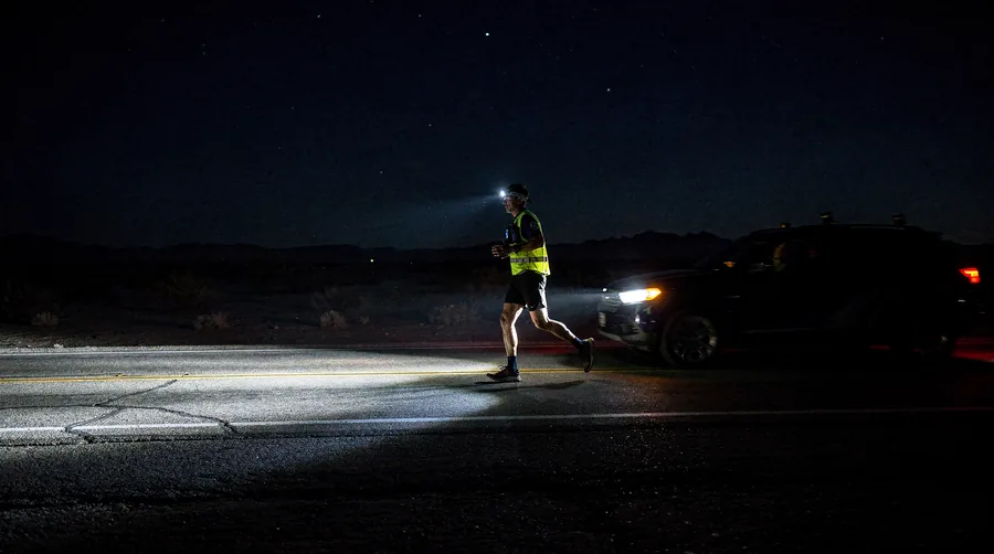 A runner in a reflective vest and headlamp on a dark highway, lit by the vehicle behind.